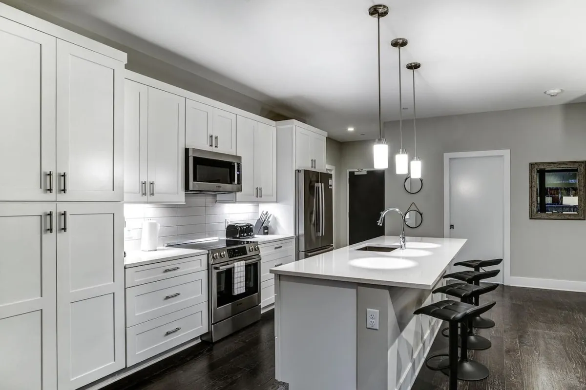 This image features a modern kitchen space with sleek cabinetry, stainless steel appliances, a spacious breakfast bar with contemporary bar stools, and stylish pendant lighting overhead. The layout clearly displays open sightlines into the adjacent living or dining area, suggesting ease of social interaction while preparing meals.