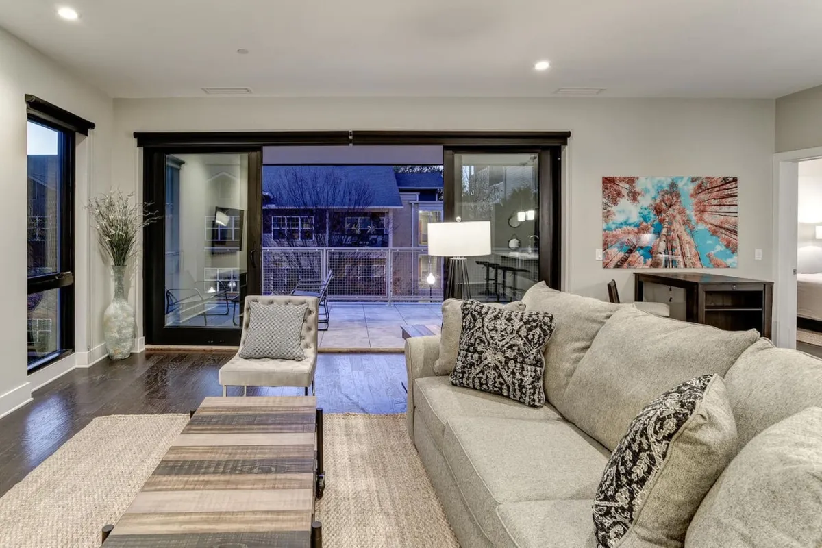 A spacious and sunlit living room with a plush sofa and coffee table is framed by expansive windows that offer breathtaking, southwest-facing views of the Blue Ridge Mountains. Vibrant artwork and thoughtfully arranged furnishings create a warm, inviting space perfect for relaxing, socializing, or soaking in mountain sunsets from the comfort of your downtown Asheville condo.