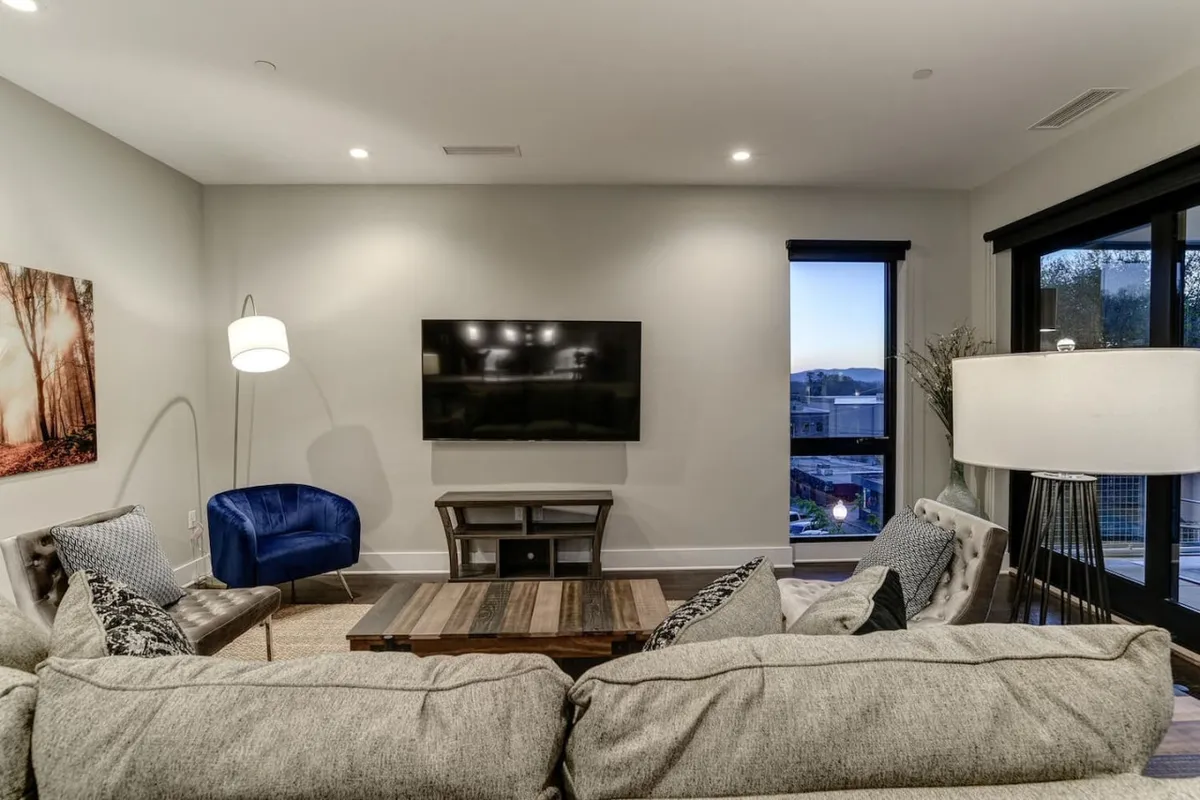 A comfortable and stylish living room with a plush sofa, blue accent chair, coffee table, television, and floor lamp. The layout is ideal for relaxed socializing, movie nights, or intimate discussions, with soft lighting creating a welcoming mood.