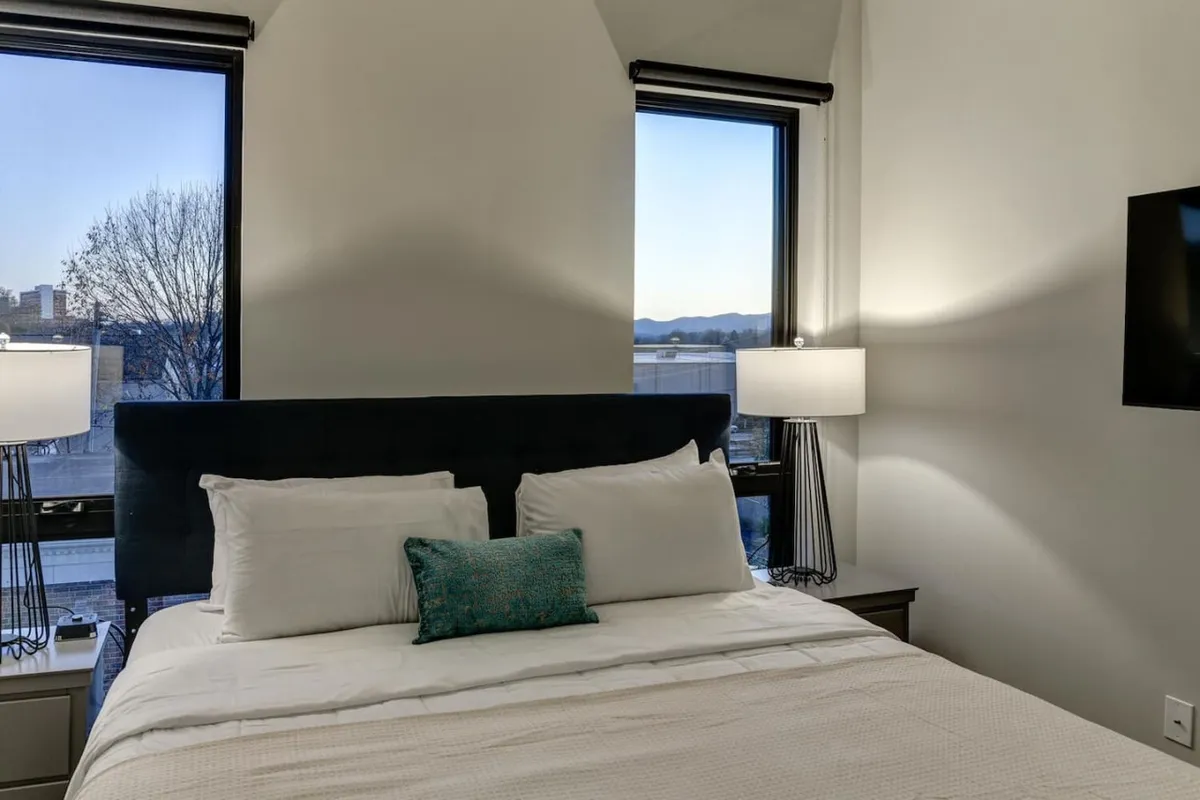 A tranquil bedroom with a comfortable bed dressed in white linens and a teal decorative pillow, flanked by windows offering a scenic mountain view. Table lamps on each side provide warm lighting, while the overall setting suggests a peaceful retreat ideal for relaxation or enjoying a book by the window.