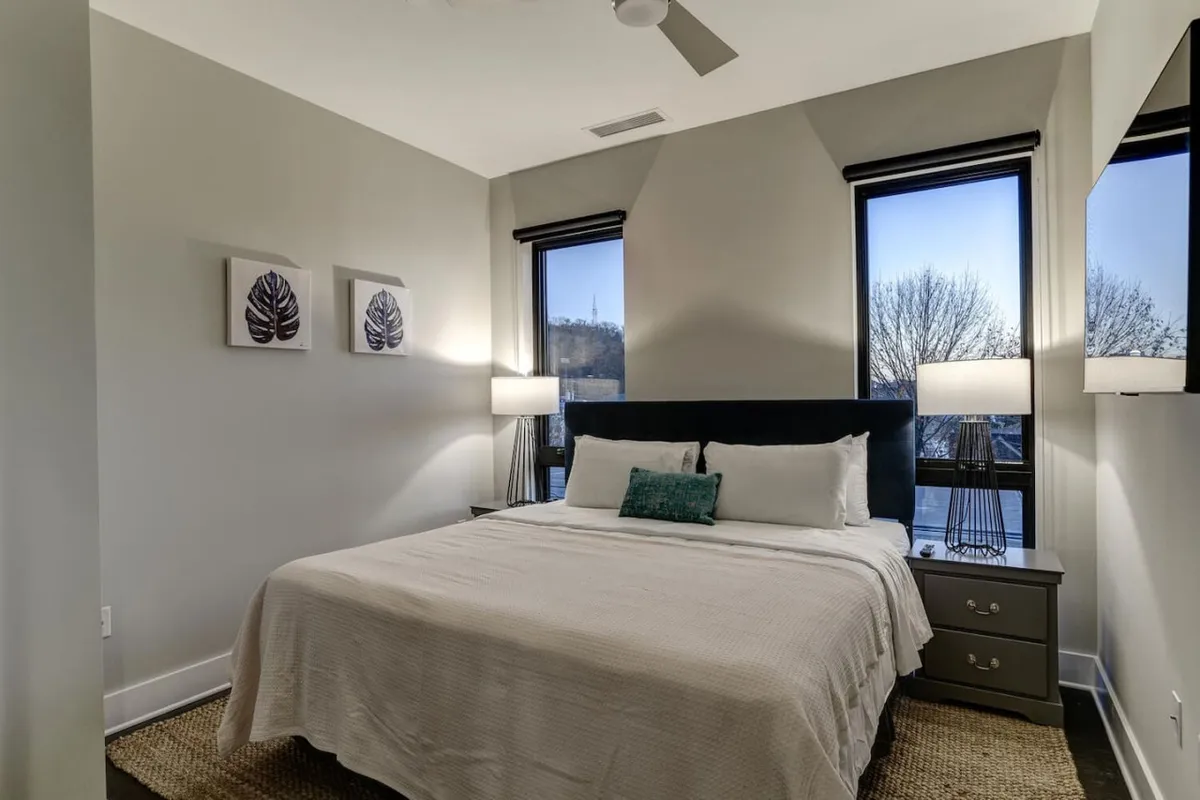 A bright bedroom featuring a large window framed by nightstands, with a plush bed and an inviting reading lamp. The room’s window offers a partial view, hinting at the world beyond and the possibility of glimpsing downtown or mountain scenery.