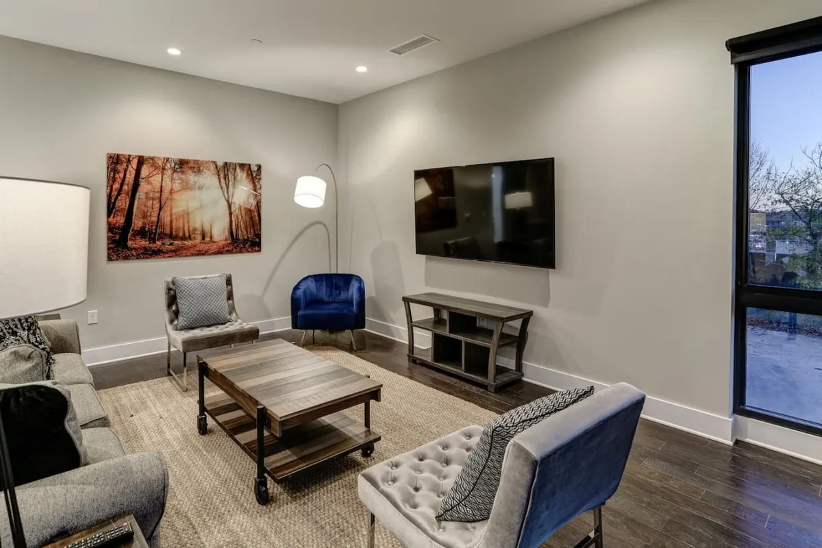 A bright, open living area featuring a plush sofa, coffee table, contemporary artwork, and a large television. Expansive windows flood the space with natural light and offer sweeping views of the Blue Ridge Mountains. The arrangement encourages conversation, relaxation, and social gatherings.