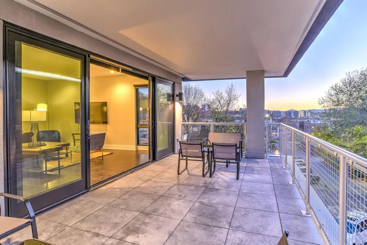 A tranquil balcony setting, featuring comfortable seating and expansive views of the sunset over the mountains. The scene includes large sliding glass doors, bridging the cozy interior with the breathtaking outdoors. The gentle evening light evokes a peaceful, contemplative mood.
