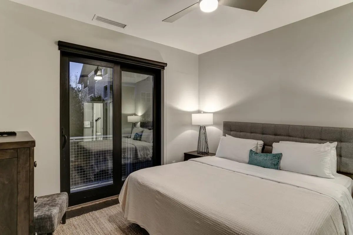 A serene bedroom featuring a plush bed with crisp white linens, a stylish glass sliding door that fills the room with natural light, a bedside table lamp, and a decorative pillow for added comfort. The calming atmosphere is perfect for rest and relaxation after a day of exploring Asheville.