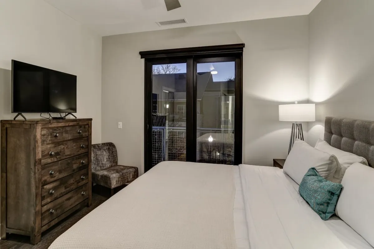 A serene king suite featuring a large bed, dresser, TV, lamp, and a sliding glass door that leads out to a private balcony or patio. The room is filled with soft morning light, suggesting a strong connection between the restful interior and the outdoor scenery.