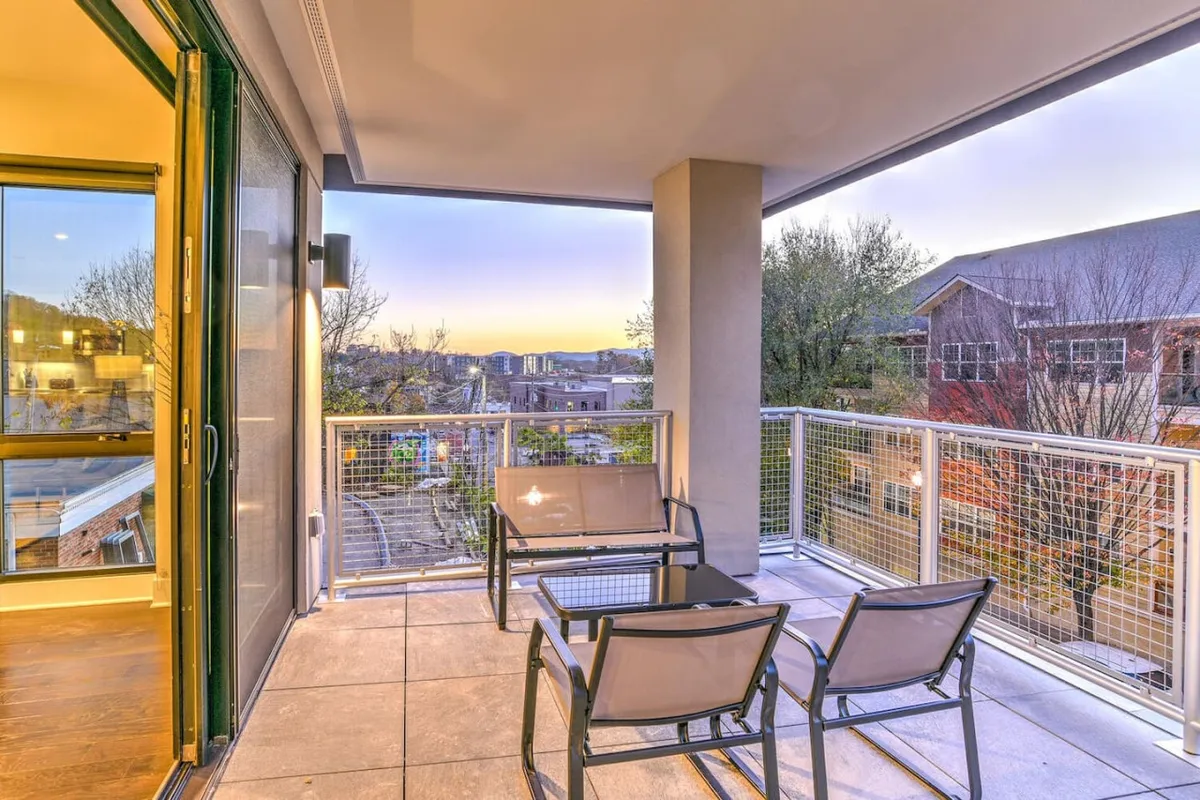 A serene, private balcony with comfortable outdoor seating overlooks sweeping Blue Ridge Mountain vistas. The space is enclosed with a sleek glass railing, offering unobstructed views of the landscape and the city lights below. The tranquil ambiance invites relaxation, reading, or enjoying the sunset.